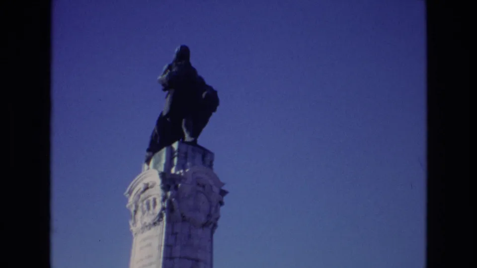 1972 Portugal 8mm frame showing the statue and upper column slightly lower in the composition, with visible film grain and saturated sky.