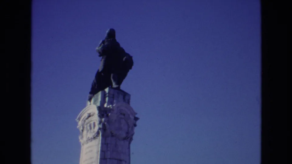 1972 Portugal home-movie frame of the same public monument, with the robed figure and column isolated against a blue sky.