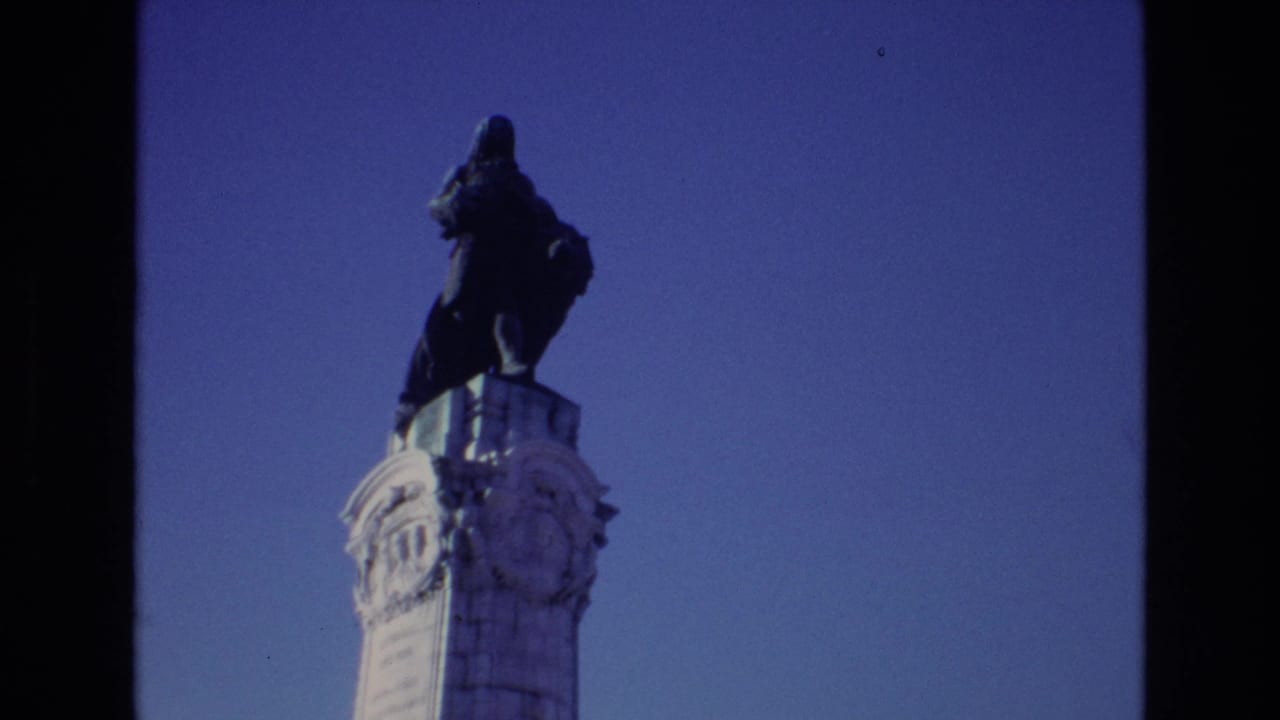 1972 Portugal 8mm frame showing a tall dark statue atop a pale ornate column against a blue sky.