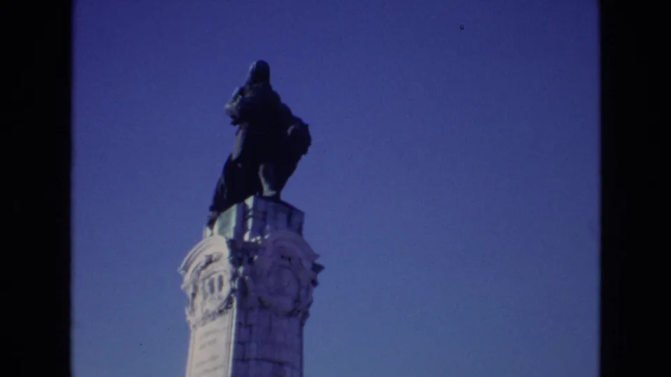 1972 Portugal 8mm frame showing a tall dark statue atop a pale ornate column against a blue sky.