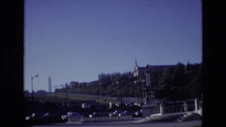 1972 Portugal 8mm frame of classic cars moving along a broad urban road with a tree-covered hill and large building beyond.