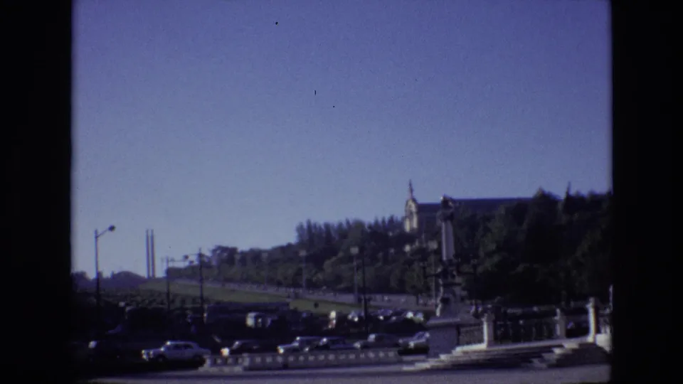 1972 Portugal 8mm frame showing moving cars on an urban roadway with trees, barriers, streetlights, and a prominent hillside building.