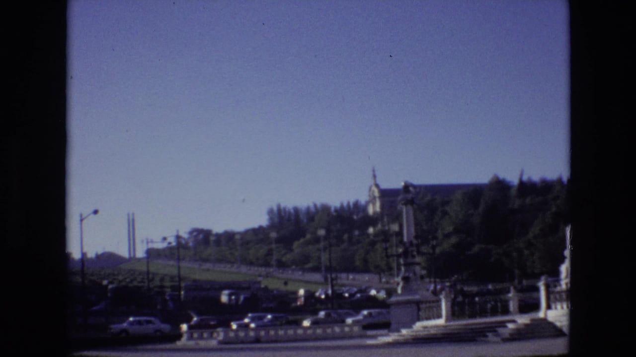 1972 Portugal 8mm frame showing a city roadway, parked cars, a stone railing, trees, and a large classical building on a hill in bright daylight.