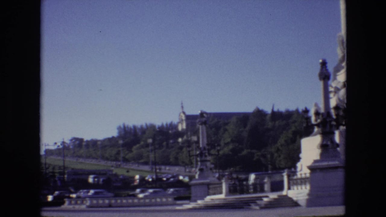 1972 Portugal home-movie frame with traffic and parked cars along a boulevard, framed by a stone balustrade, greenery, and a distant civic building.