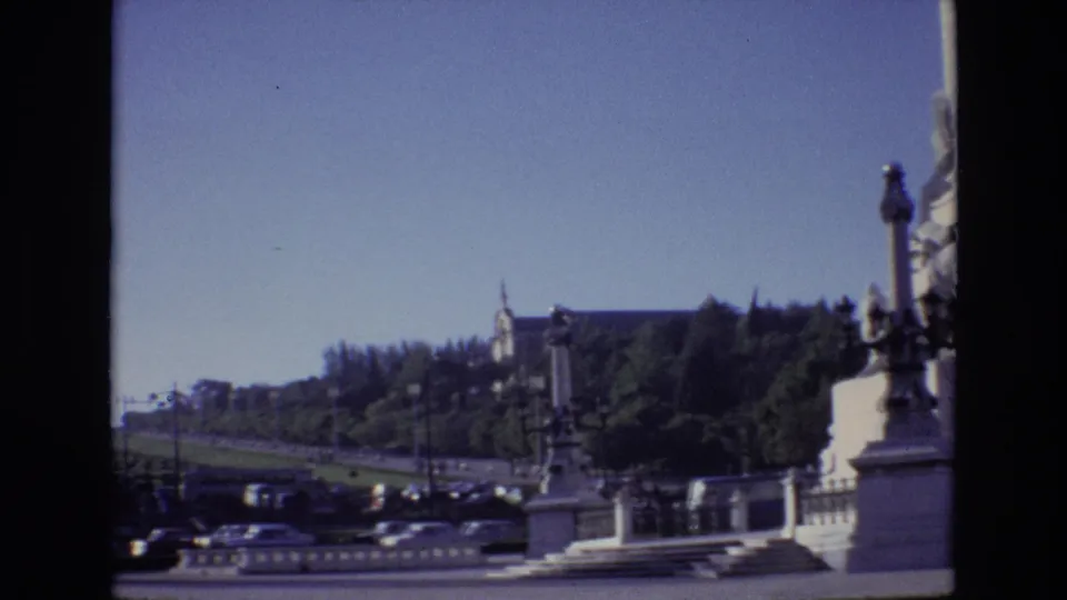 1972 Portugal home-movie frame with traffic and parked cars along a boulevard, framed by a stone balustrade, greenery, and a distant civic building.