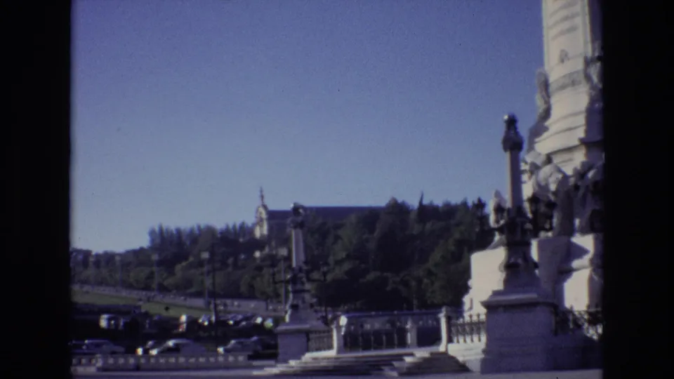 1972 Portugal 8mm frame from near a stone monument, looking across a boulevard with cars toward trees and a large building on the hillside.