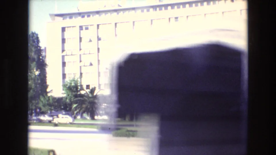 1972 Portugal home-movie frame showing a sunlit building facade, palm planting, cars near the street, and a blurred foreground object partly blocking the view.