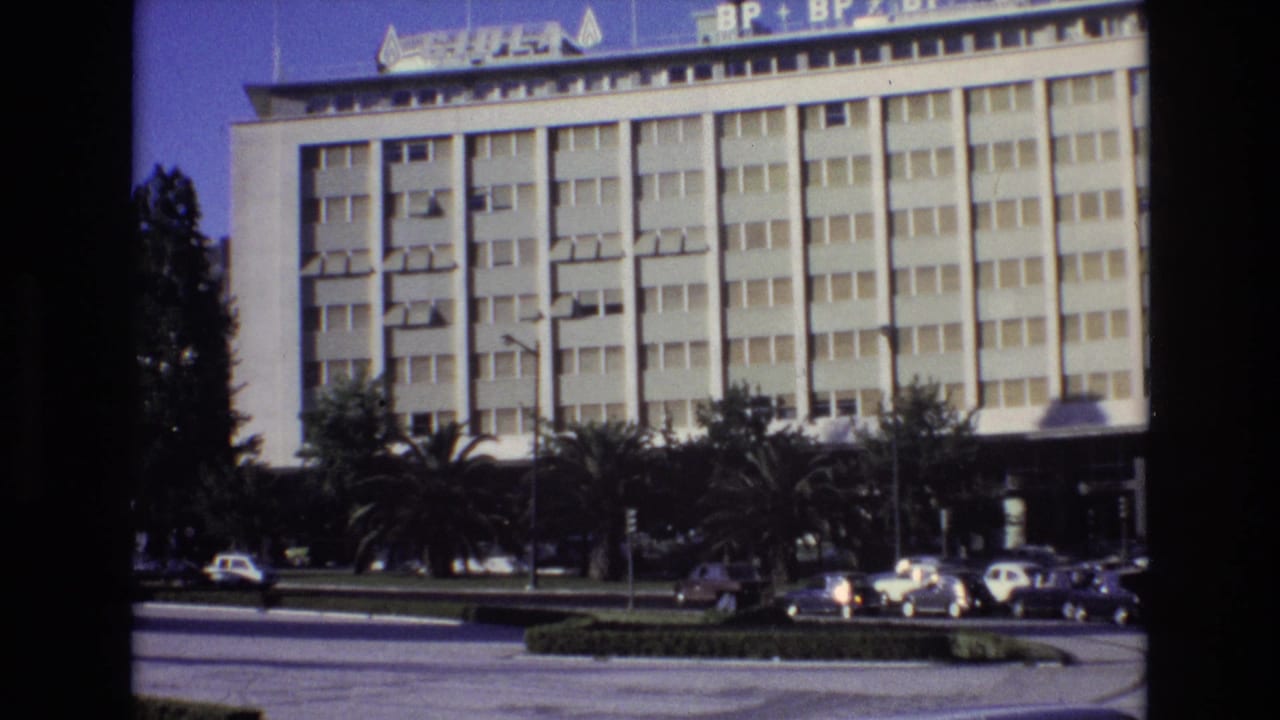 1972 Portugal 8mm frame showing a large white gridded building with rooftop signage, palm trees, a planted median, and cars moving along the road.