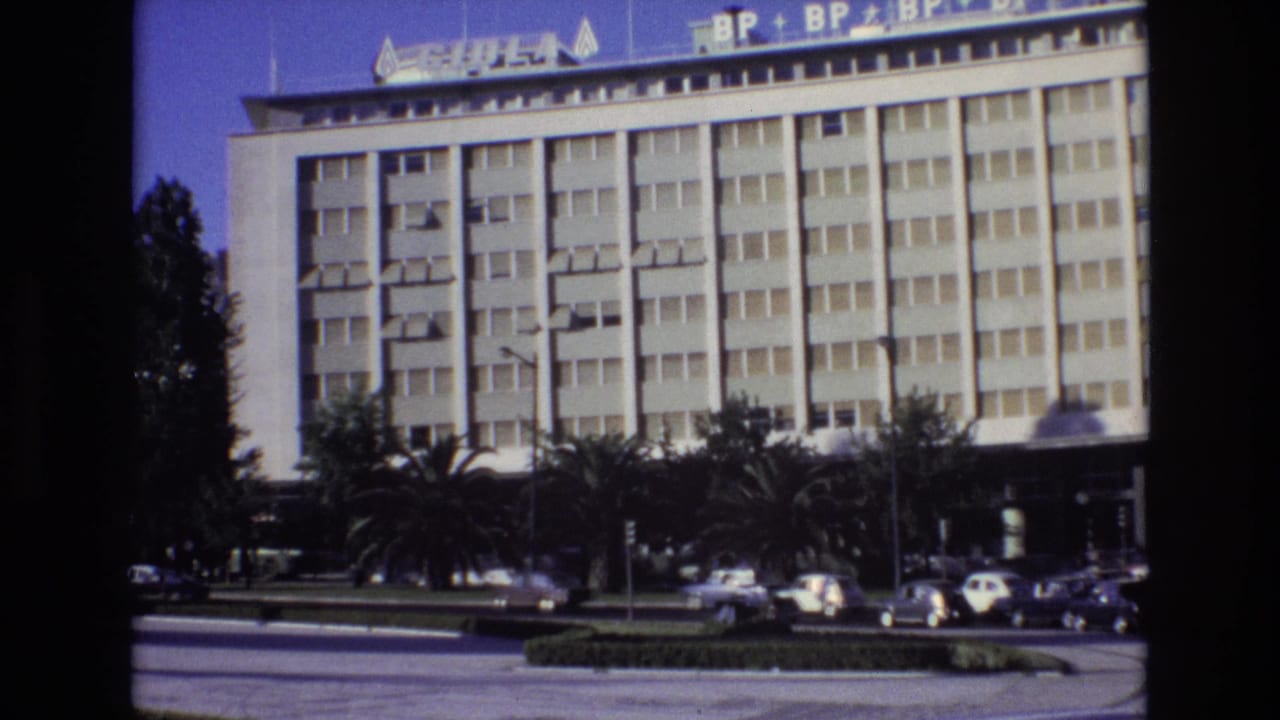 1972 Portugal street view with a white modern building, visible rooftop commercial signage, roadside trees, palms, and several cars passing in front.