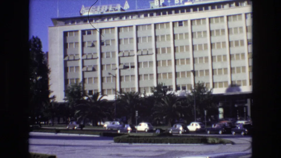 1972 Portugal 8mm frame holding on the white gridded facade while classic cars pass near palm trees and a low planted median.