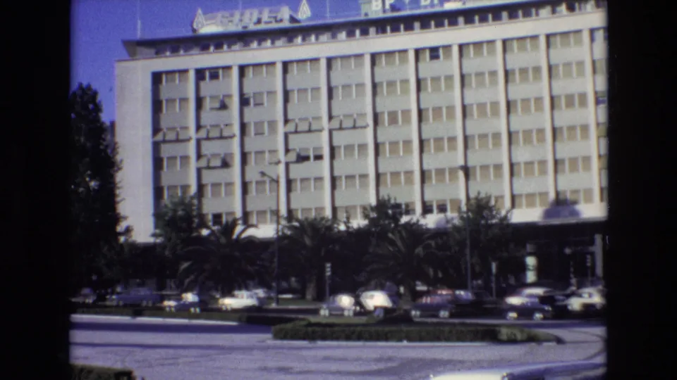 1972 Portugal street frame with a white multi-story building, rooftop signage, palms, a curved roadway, and multiple cars in front.