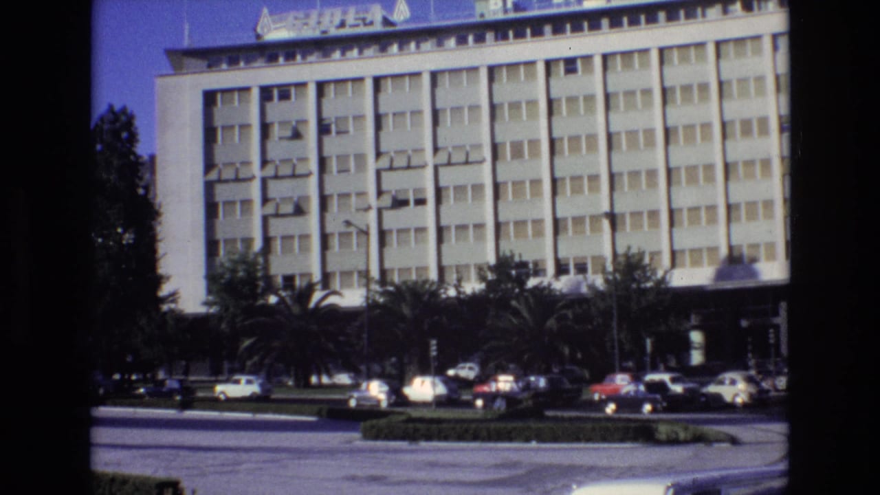 1972 Portugal home-movie frame showing a modern multi-story city building, palm trees, and classic cars moving or parked along a broad roadway.