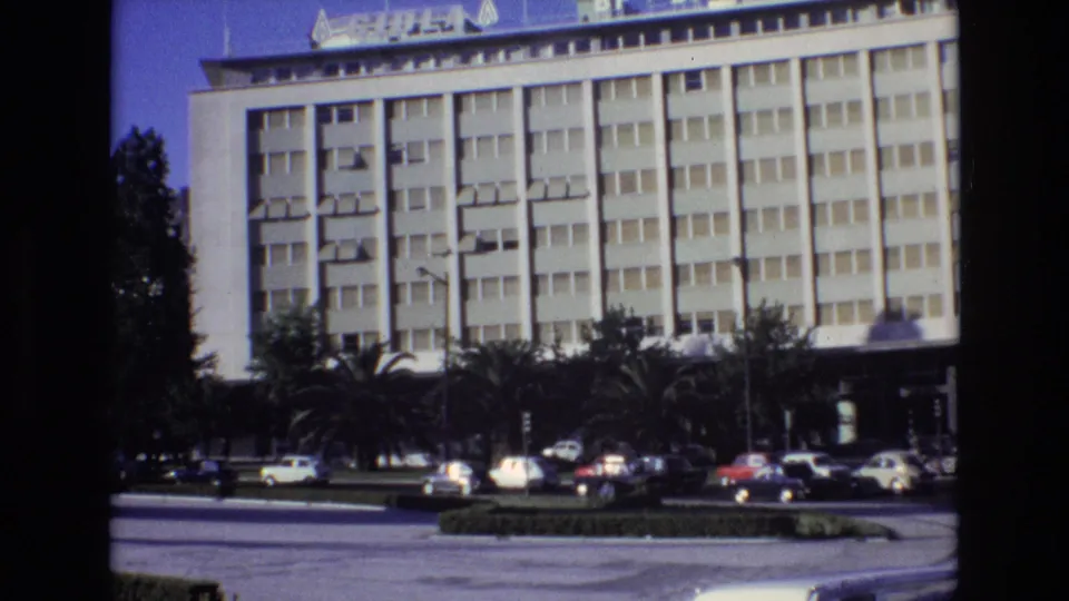 1972 Portugal home-movie frame showing a modern multi-story city building, palm trees, and classic cars moving or parked along a broad roadway.