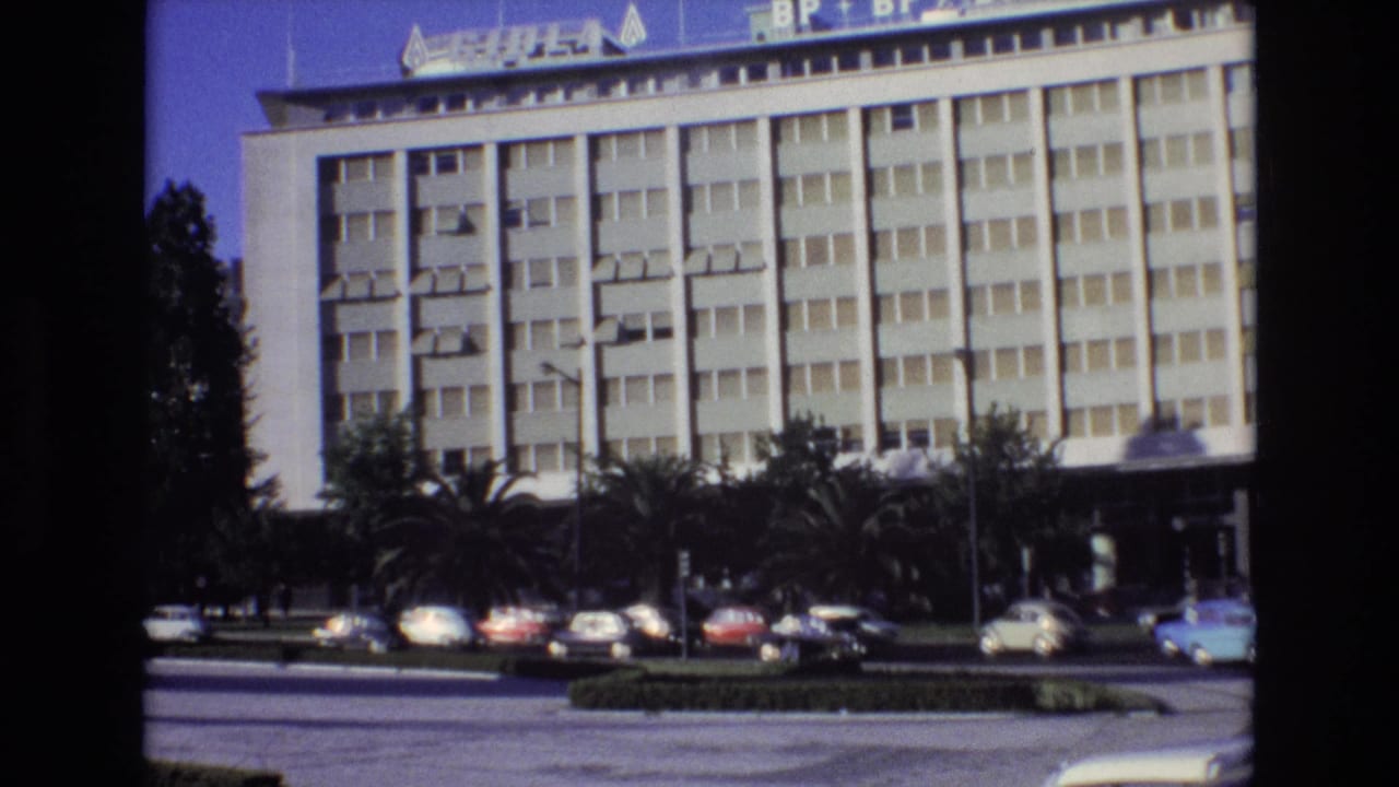 1972 Portugal street view with a large modern building, rooftop commercial signs, palm trees, and early-1970s cars along the curb and roadway.