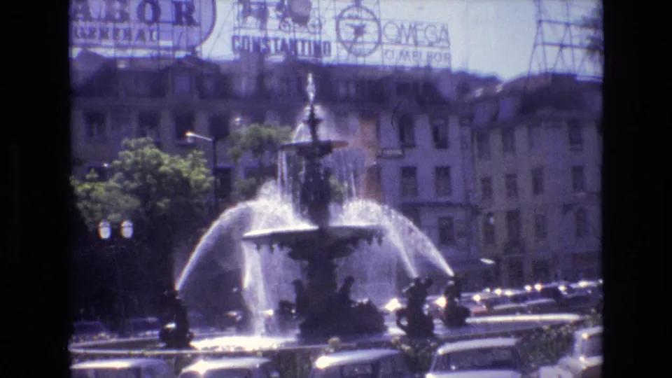 1972 Portugal home-movie view of a public fountain spraying water in front of traffic, trees, older building facades, and partially visible commercial signage.