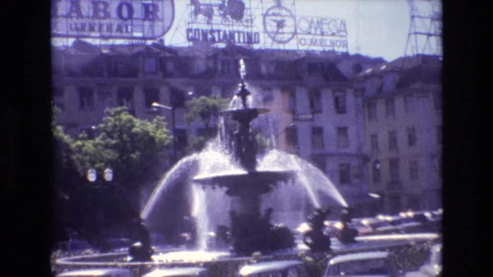 1972 Portugal frame showing a central fountain, moving cars, trees, street lights, and rooftop commercial signs above surrounding city buildings.