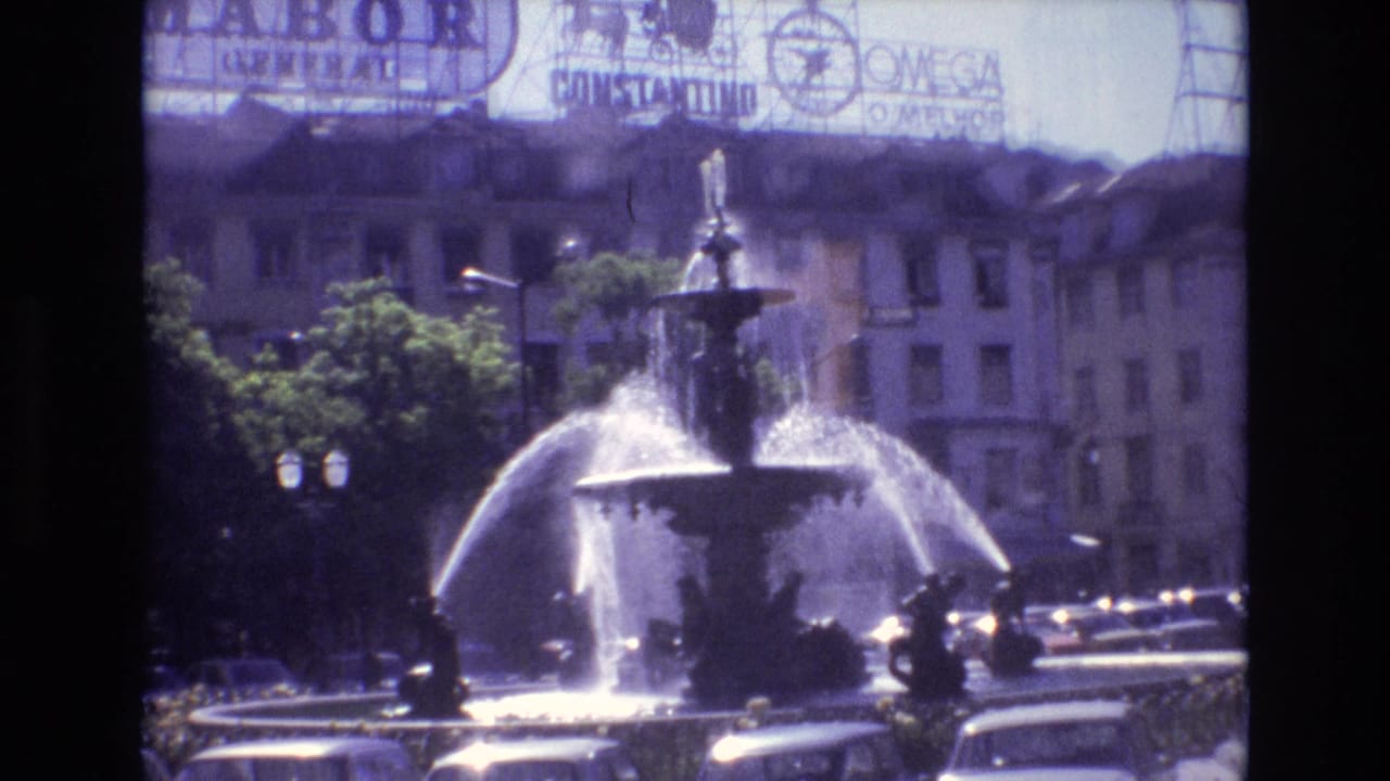 Portugal 1972 8mm frame of a large public fountain spraying water, with early-1970s cars, trees, buildings, and visible rooftop advertising signs in the background.