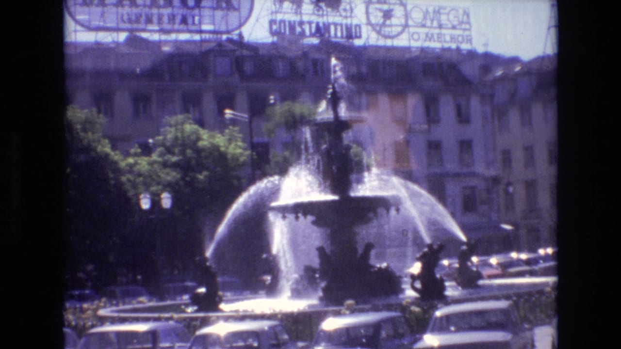 Fountain spraying in front of trees, parked cars, street lamps, buildings, and rooftop advertisements.