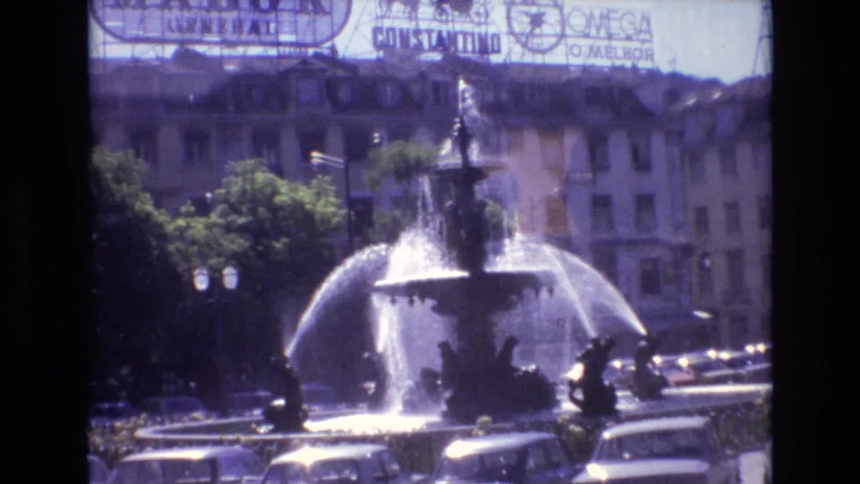 Portugal 1972 8mm frame of the fountain from a steady visitor viewpoint, with bright spray, shaded trees, cars, street lamps, and billboard signage behind it.