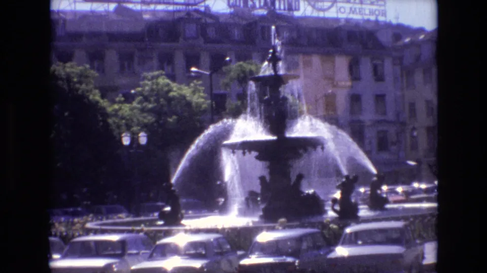Portugal 1972 frame showing the fountain at the end of the clip, with sunlit water, cars clustered around the square, trees, street lamps, and commercial rooftop signage.