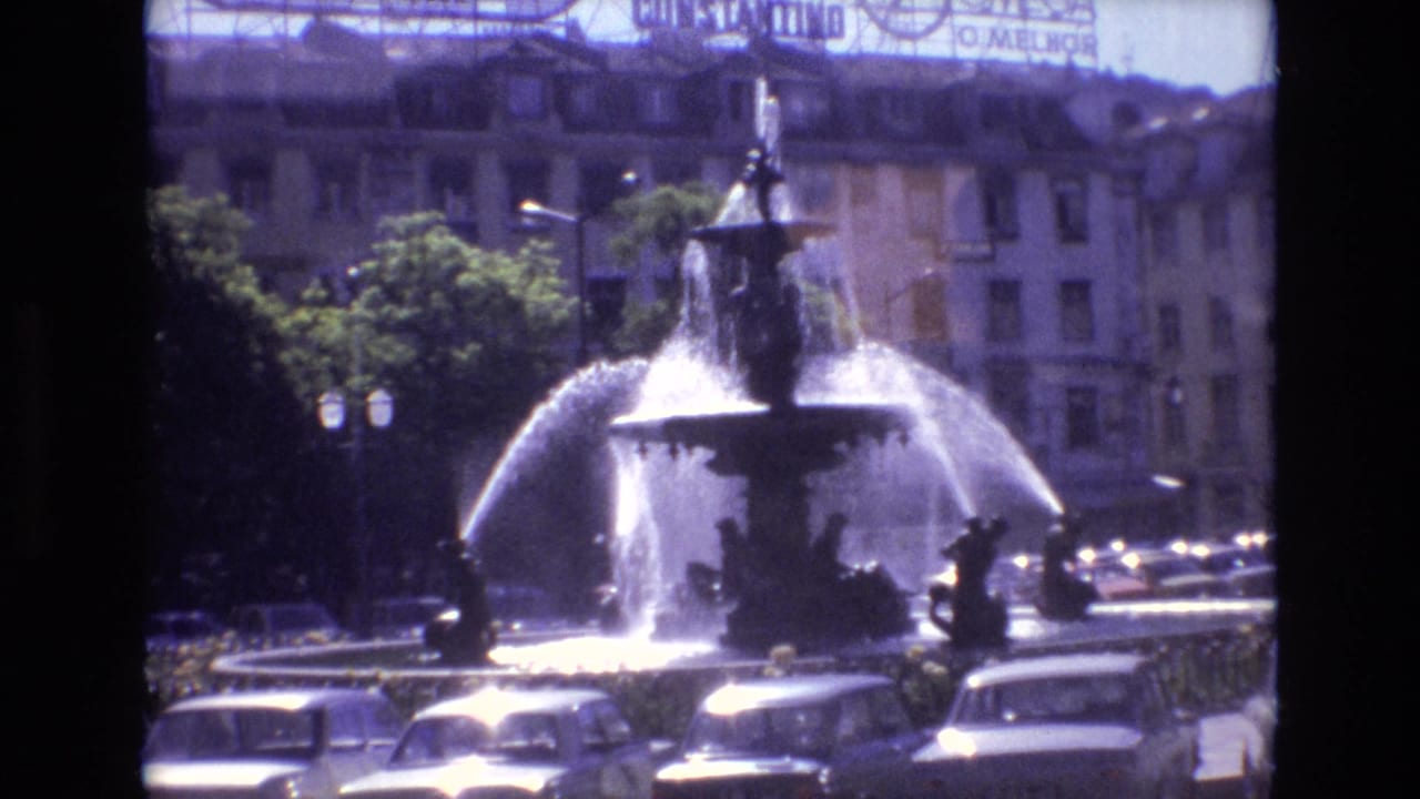 Large city fountain spraying water with cars and buildings behind it.
