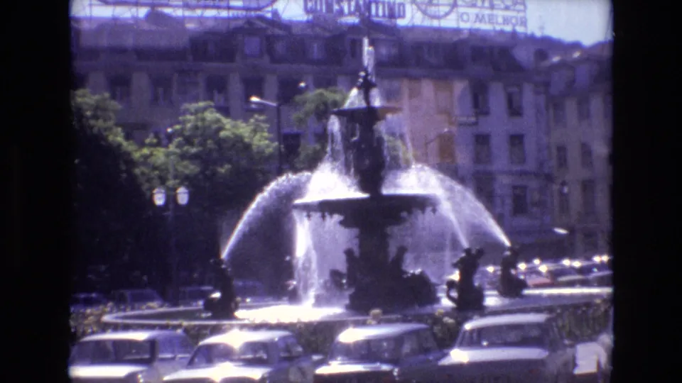 1972 Portugal 8mm frame showing an ornate urban fountain spraying in multiple arcs, with cars in the foreground and building facades with signage behind it.