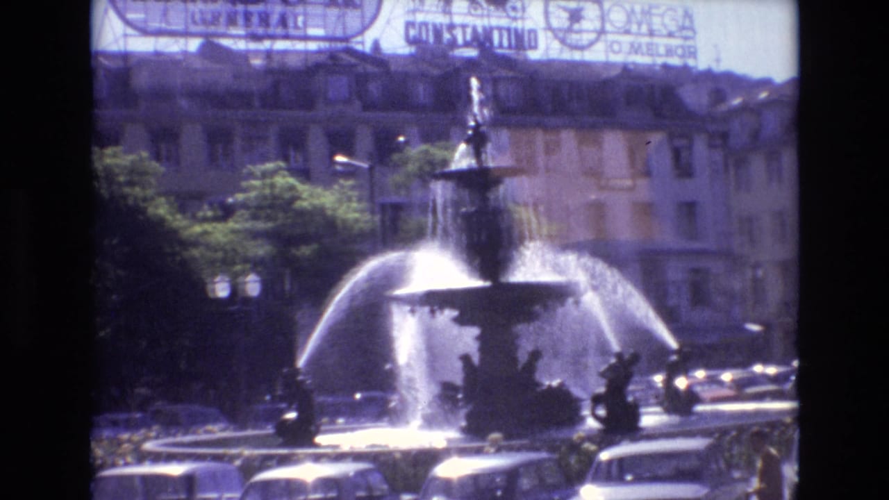 Fountain spraying across a city square with cars and background signage.