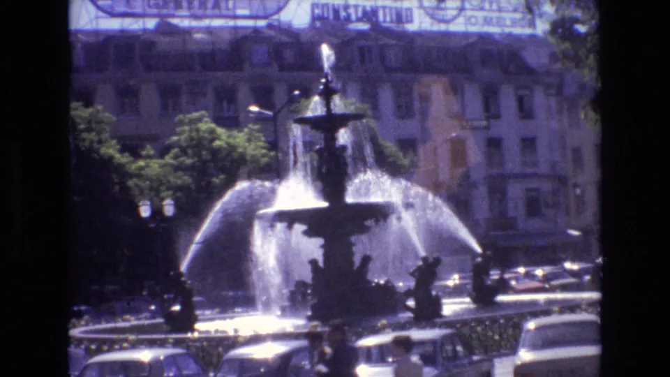 1972 Portugal home-movie frame of an ornate public fountain, bright water spray, roadside cars, trees, and tall urban buildings in the background.