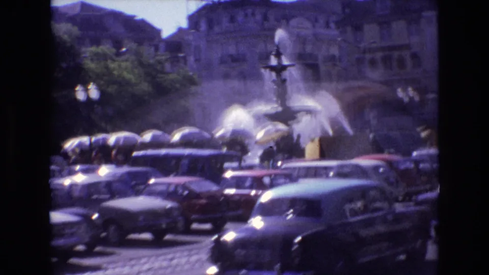 A wider 1972 Portugal city-square view shows the fountain partly obscured by passing and parked cars, with pedestrians near the basin and buildings beyond.