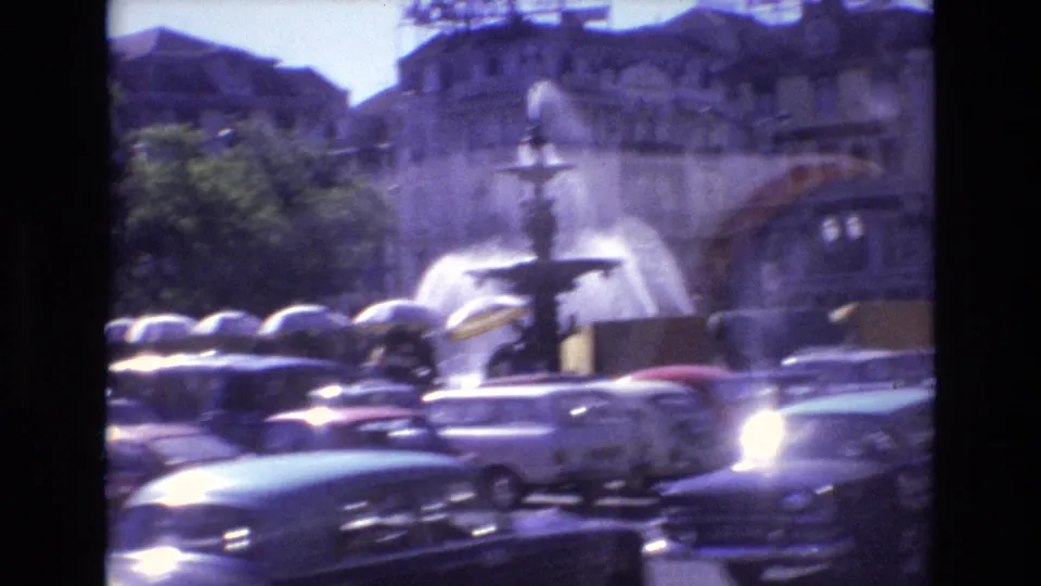 1972 Portugal 8mm frame showing a tiered public fountain spraying water behind classic cars and distant pedestrians in a city square.
