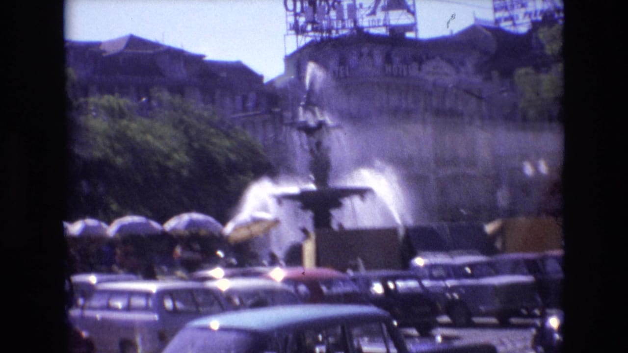 1972 Portugal 8mm frame with classic cars in the foreground, a large fountain spraying water, and pedestrians gathered under umbrellas near trees.