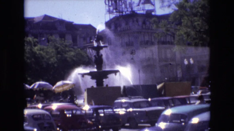 1972 Portugal 8mm frame centered on the fountain, with water spray rising above cars and distant pedestrians in a sunlit urban square.