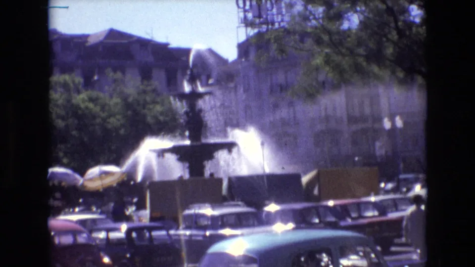 1972 Portugal 8mm frame showing the public fountain and water spray above a line of classic cars, with trees and building facades beyond.