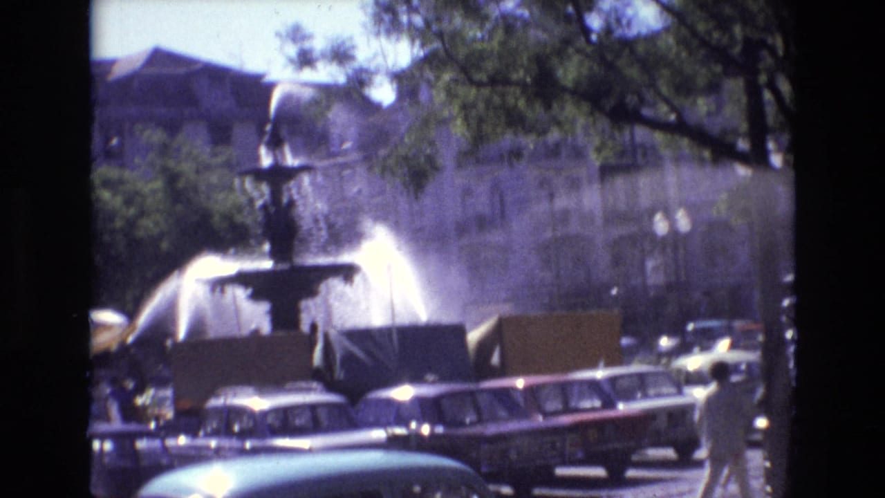 Fountain spraying water beside parked cars and trees in an urban square.