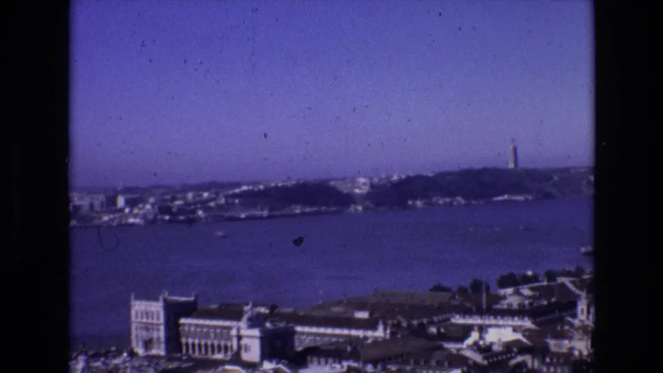 1972 Portugal 8mm frame showing a wide coastal bay, waterfront buildings, distant hills, and a tall tower on the horizon.