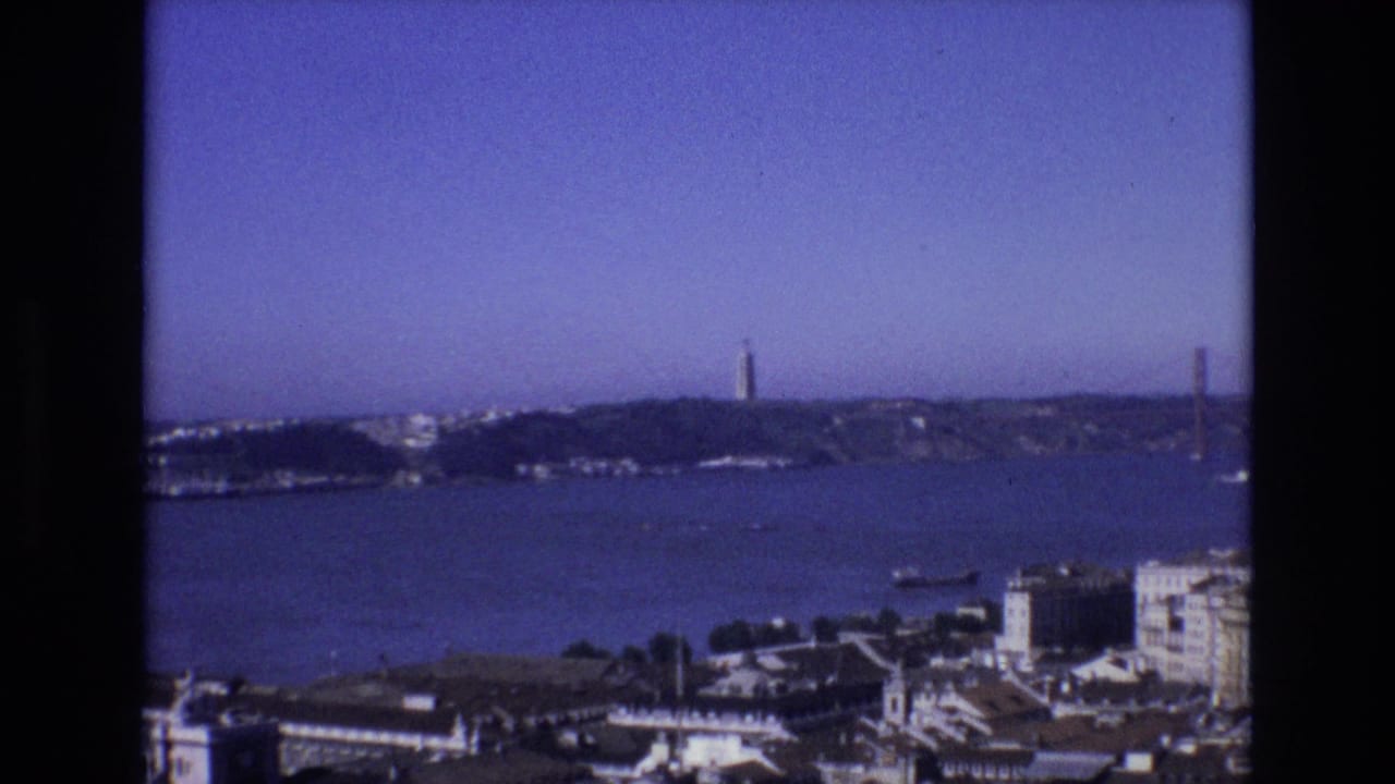1972 8mm home-movie frame looking across Lisbon and the Tagus River, with Torre de Belem and the bridge visible beyond waterfront buildings.