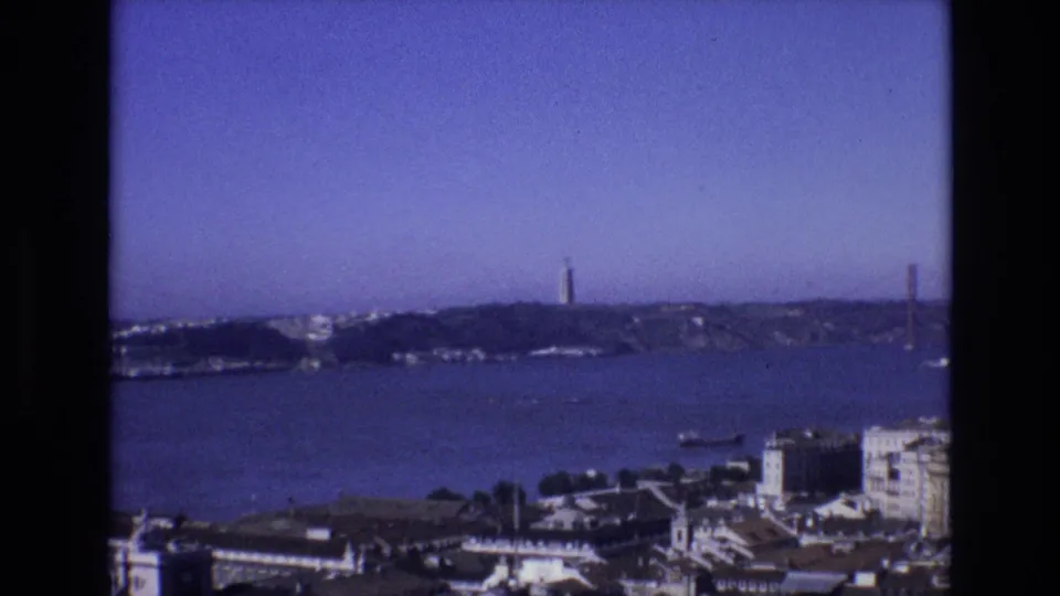 1972 8mm home-movie frame looking across Lisbon and the Tagus River, with Torre de Belem and the bridge visible beyond waterfront buildings.