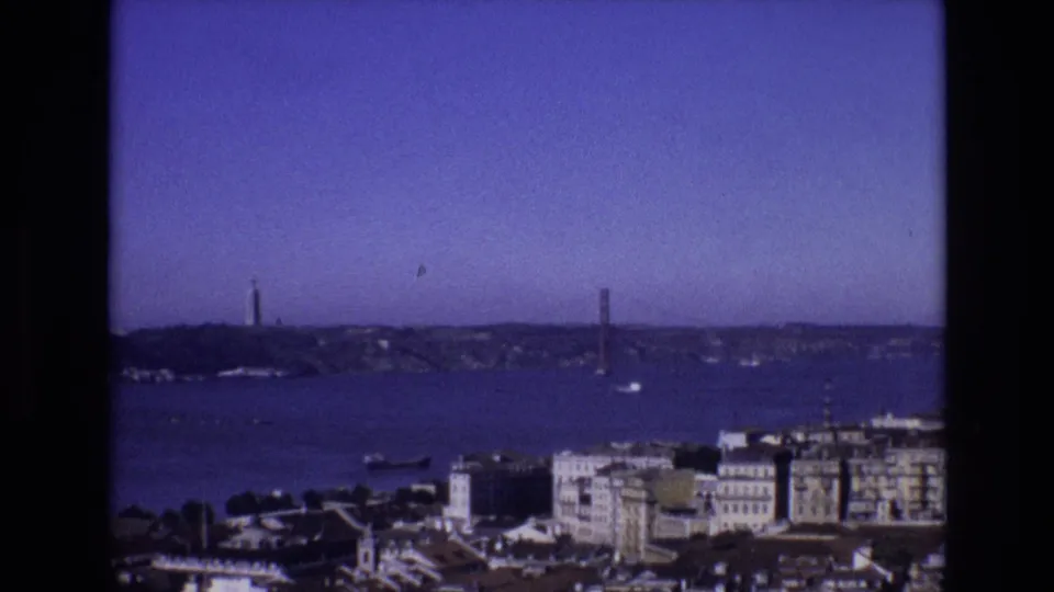 1972 Lisbon travel footage frame showing foreground city rooftops, the Tagus River, Torre de Belem, and the suspension bridge under a clear blue sky.
