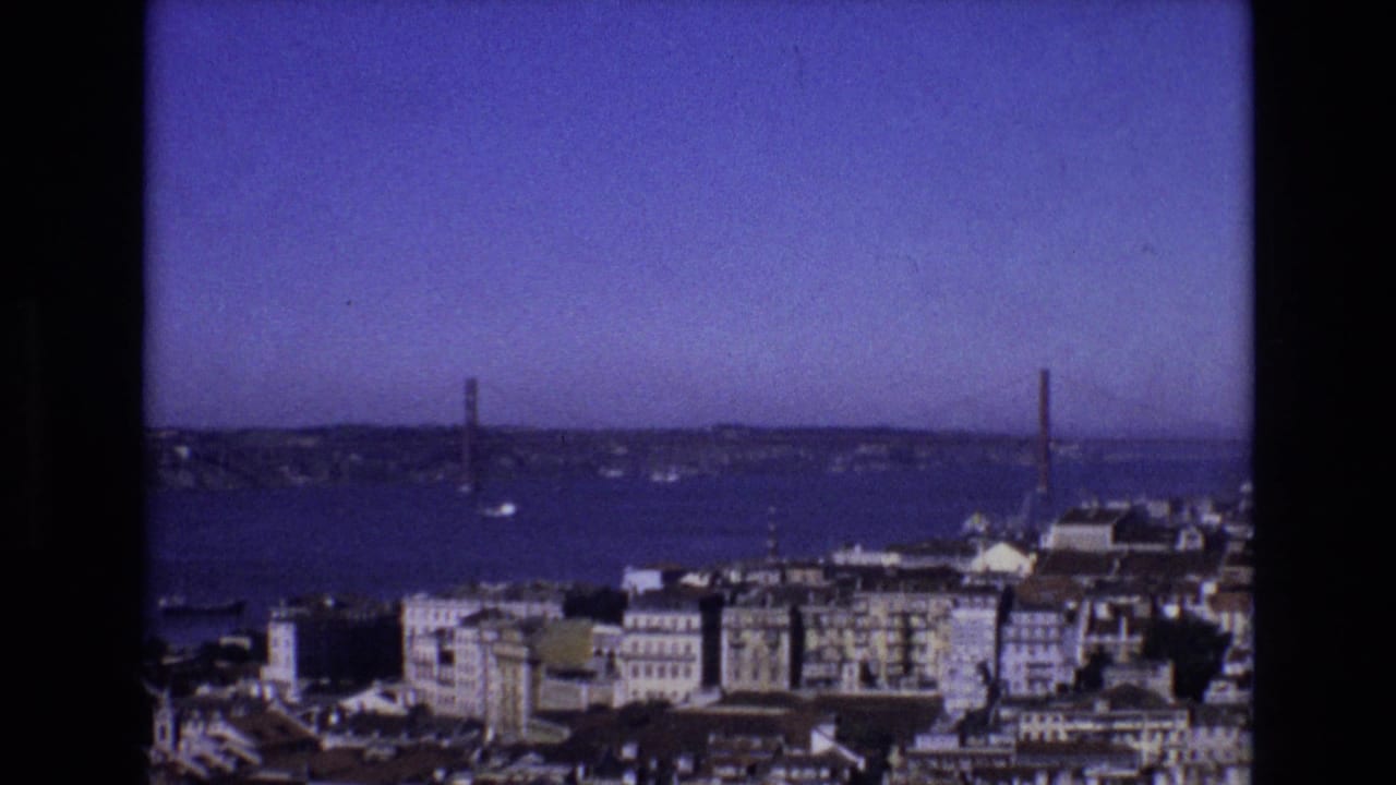 1972 Portugal home-movie frame showing Lisbon rooftops and waterfront buildings below the 25 de Abril Bridge spanning the Tagus River.