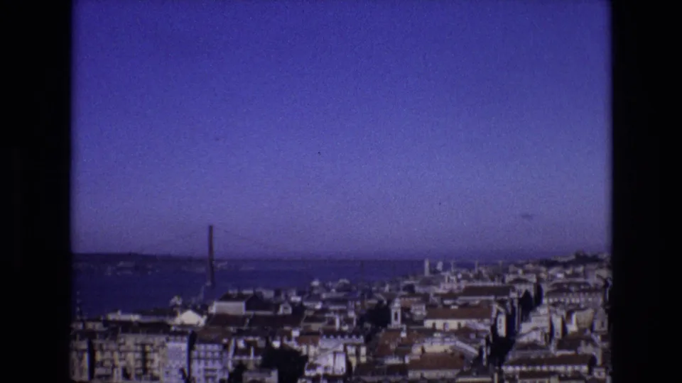 1972 Portugal 8mm frame showing an elevated view across city rooftops toward a wide river or harbor and a distant suspension bridge.