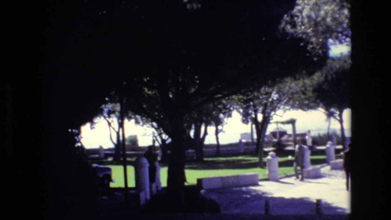 1972 Portugal 8mm frame of a sunlit public promenade partly shaded by trees, with pedestrians near benches and white pillars overlooking the water.