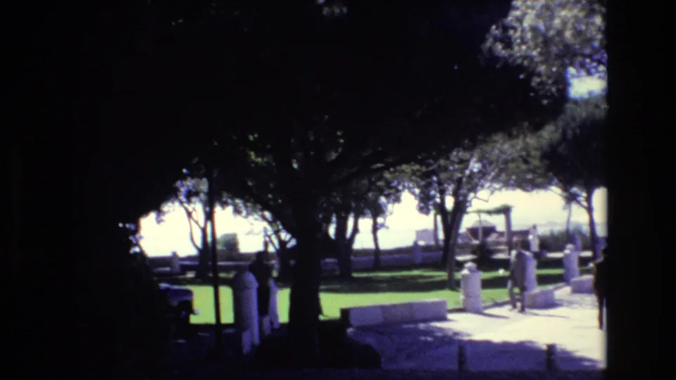 1972 Portugal 8mm frame of a sunlit public promenade partly shaded by trees, with pedestrians near benches and white pillars overlooking the water.