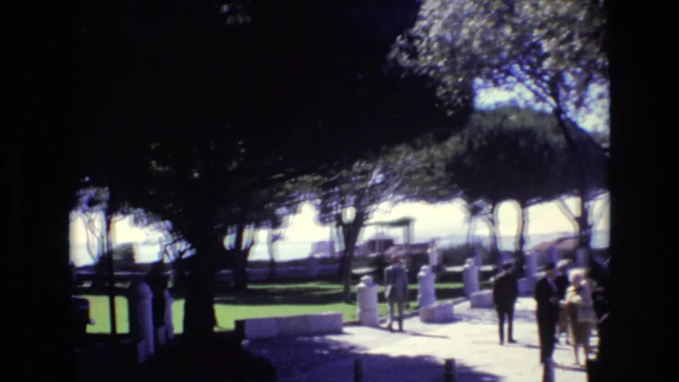 1972 Portugal 8mm frame showing adults walking and standing along a bright paved promenade beneath trees, with benches and white posts nearby.