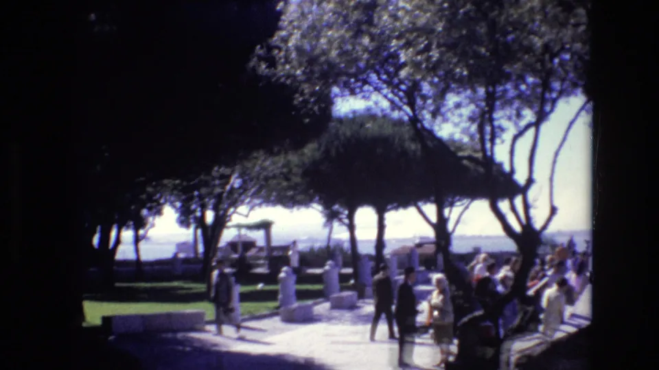 1972 Portugal 8mm frame of several adults gathered or walking on a tree-lined public promenade, with benches, white posts, and a waterfront view in the background.