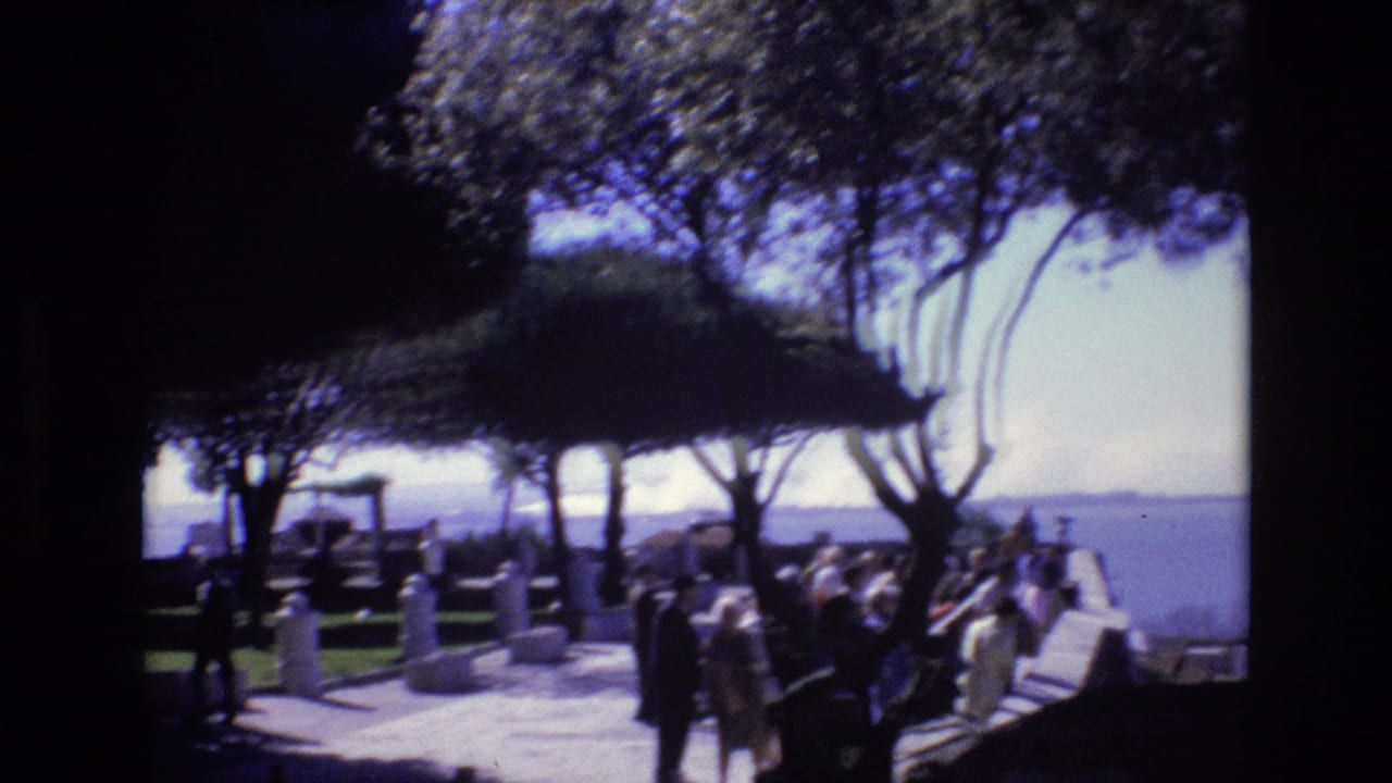1972 Portugal home-movie frame showing visitors in light clothing walking through a shaded park or zoological center with water visible beyond the trees.