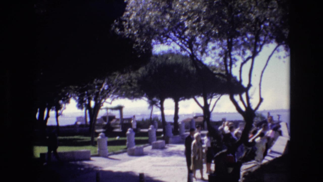 1972 Portugal frame with visitors gathered along a sunlit path in a landscaped zoological or park setting, with trees and water in the background.