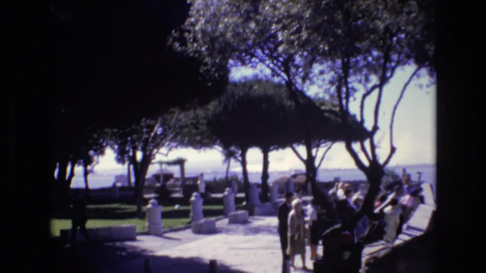 1972 Portugal frame with visitors gathered along a sunlit path in a landscaped zoological or park setting, with trees and water in the background.
