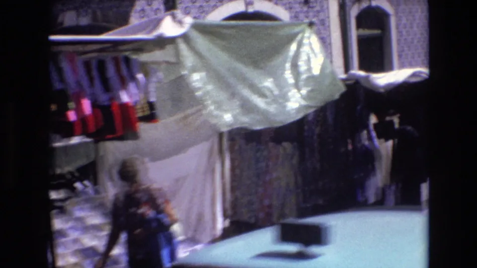 A 1972 Portugal street-market view shows hanging merchandise under a pale awning against a blue-and-white tiled facade, with a blurred adult passerby near the stall.