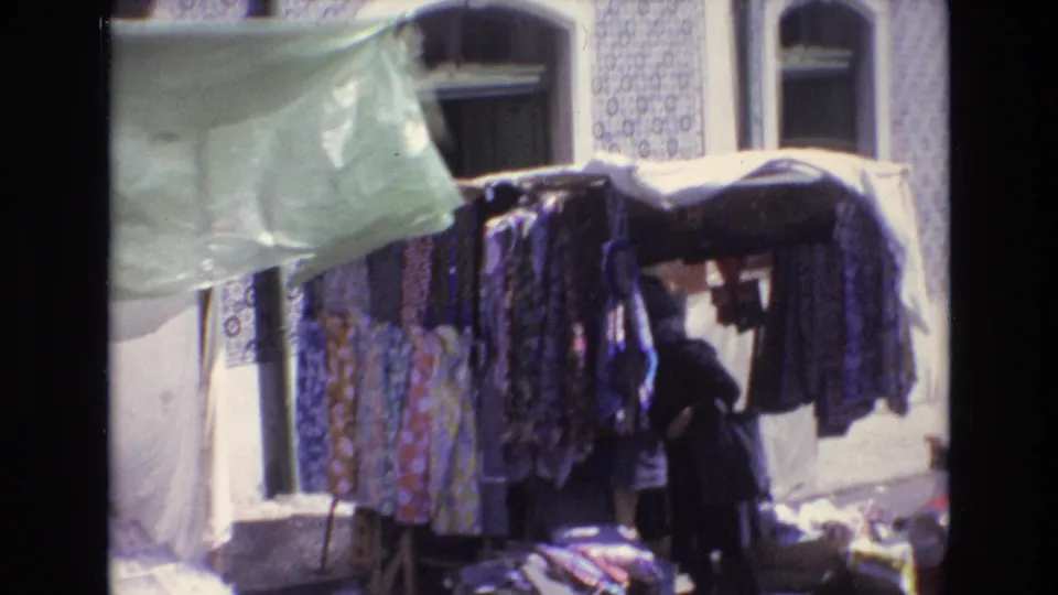 A 1972 Portugal market frame shows patterned fabrics and garments hanging from a stall outside a tiled building, with an adult shopper partly visible below the awning.