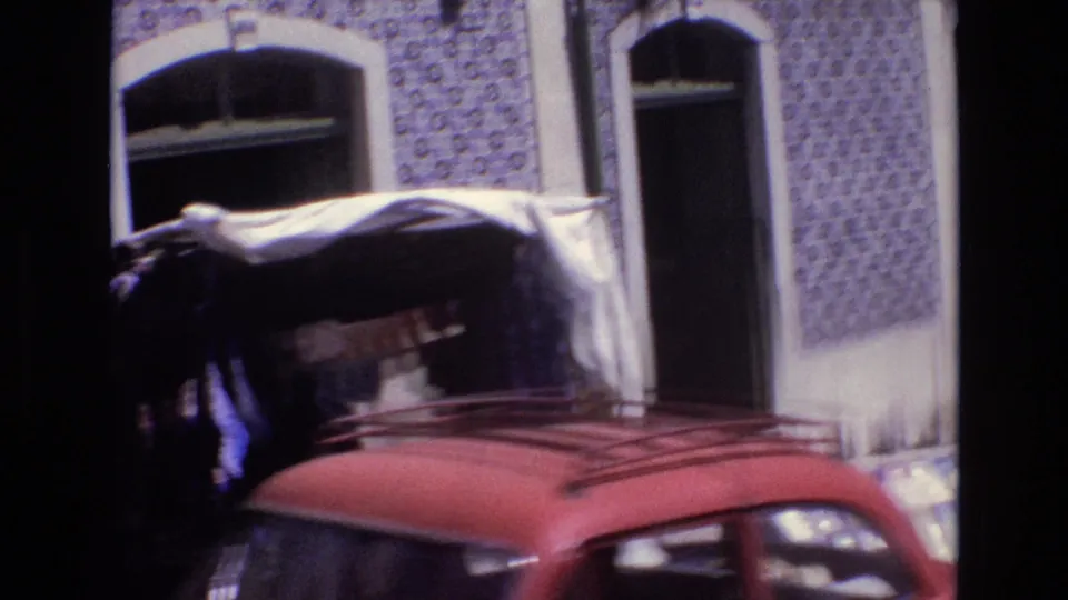 A 1972 Portugal home-movie frame shows a blue-and-white tiled facade with arched openings, market awnings, hanging textiles, and the roof of a red car in the foreground.